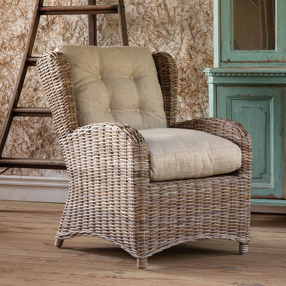 A wicker armchair with a tufted beige back cushion and a light beige seat cushion, set in a rustic room with a wooden ladder, distressed wallpaper, and a teal cabinet.