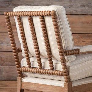 A close-up of a wooden armchair with cream-colored striped fabric cushions and a natural wood spindle-back frame, set against a rustic wood plank wall.