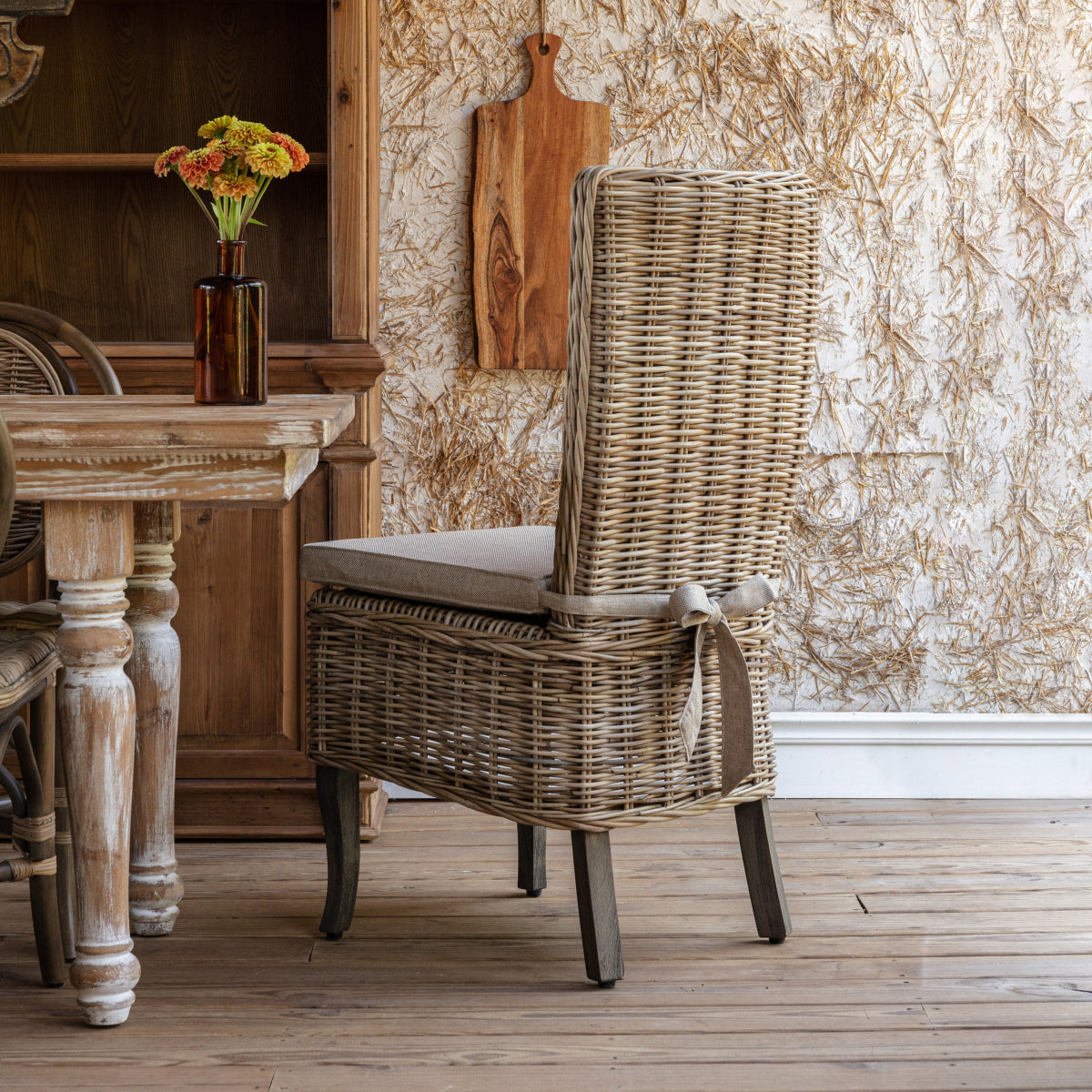 Copperfield Rattan Dining Chair with a tall woven back and linen cushion beside a rustic dining table