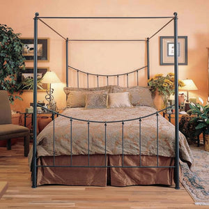 A black metal four-poster canopy bed with a curved headboard and footboard, styled in a warm, rustic bedroom setting with peach walls, wooden floors, and decorative plants.